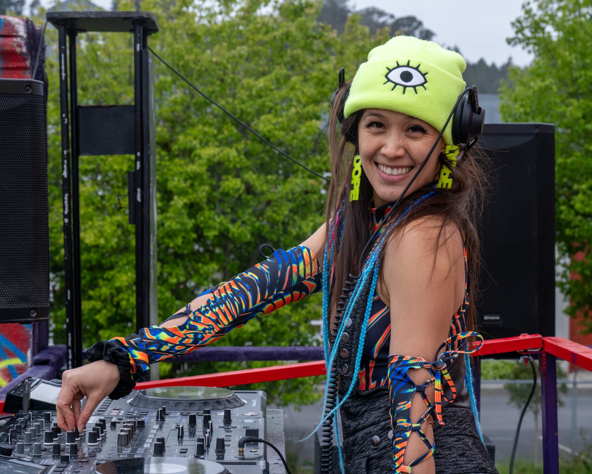Aster smiling at the decks in a neon beanie and headphones during an outdoor set.