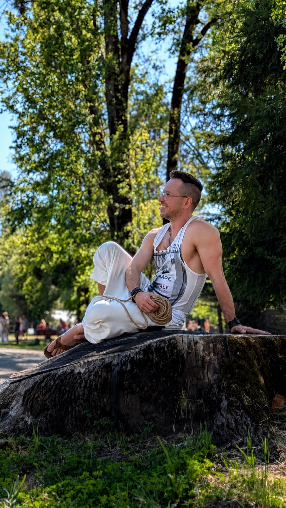Davide sitting on a tree stump outdoors with a coil of rope in his lap, looking off to one side.
